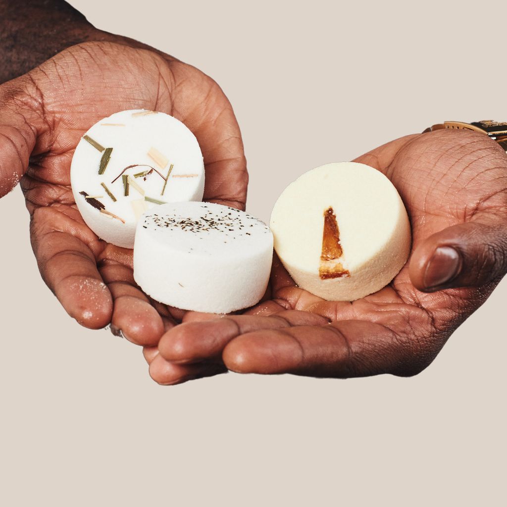 Man holding three handmade aromatherapy shower steamers, each topped with unique botanicals. The first steamer, 'Clarity,' is infused with lemongrass, rosemary, and tea tree. The second, 'Vitality,' features orange, lemon, and lime. The third, 'Purity,' includes eucalyptus, peppermint, and lemon.